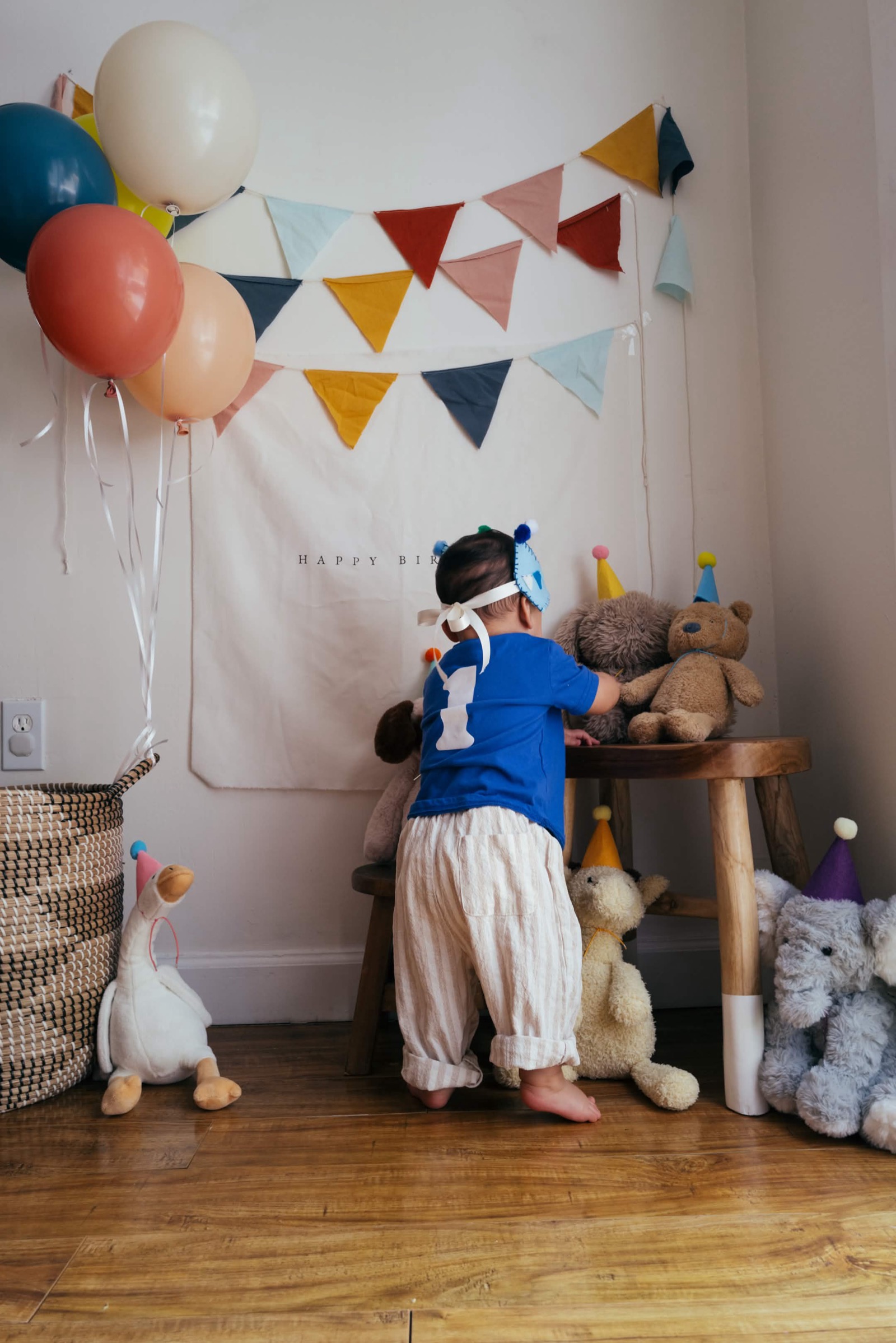 Roman in a blue '1' shirt and party hat, reaching toward stuffed animals under a HAPPY BIRTHDAY banner and bunting, balloons in the corner.